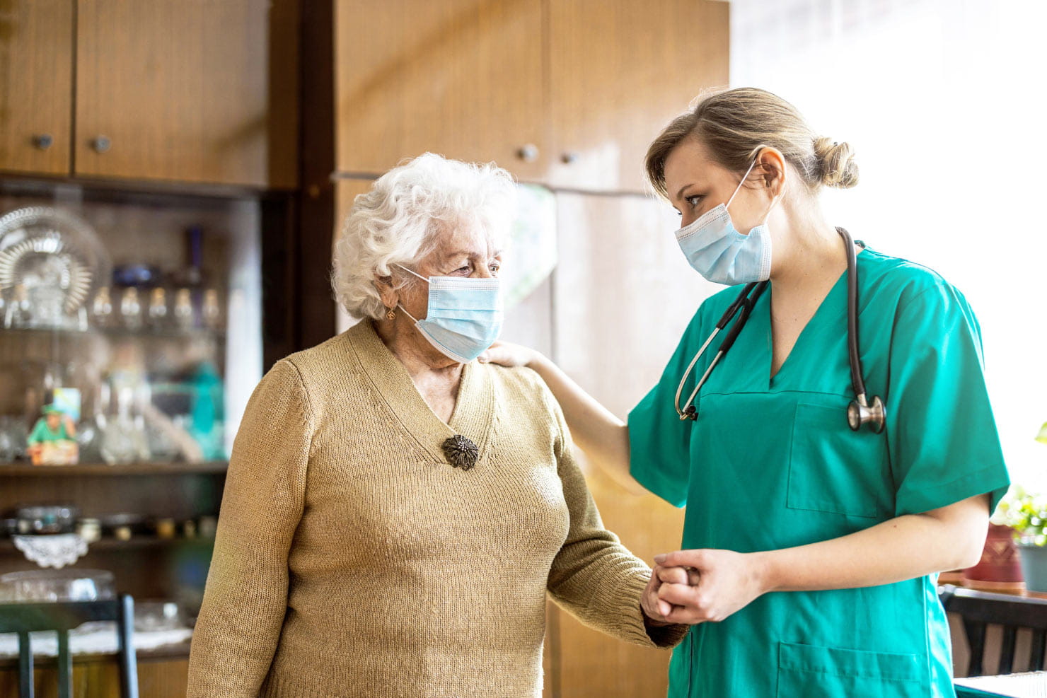 A masked female nurse stands next to a masked elderly patient while holding her hand